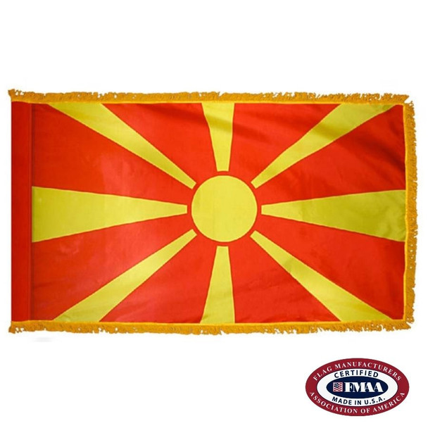 The indoor Macedonia flag has 3 sides of gold fringe around a red field with a yellow sun in the center and 8 long rays. FMAA Certified Made in America seal.