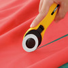 Close-up of a hand using the OLFA 45mm Quick-Change Rotary Cutter to cut through red fabric, showing the cutter's blade and ergonomic yellow handle.