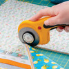 Close-up of a hand using the OLFA Deluxe Ergonomic mm Rotary Cutter to cut through multiple layers of fabric, showing the cutter's blade and ergonomic yellow handle.