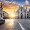 Sunrise over the Piazza del Duomo in Milan, with the sun casting a warm glow on the Gothic facade of the Duomo di Milano and the surrounding buildings, the square quiet and empty.