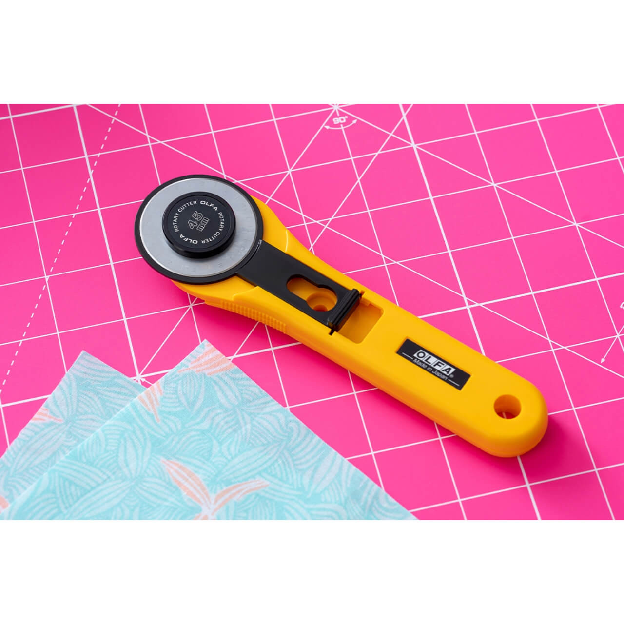 OLFA rotary cutter with a yellow handle and black circular blade on a pink OLFA cutting mat, alongside a piece of patterned fabric, ready for precision crafting.