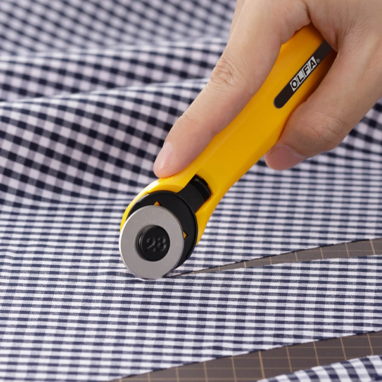 Close-up of a hand using the OLFA 28mm Quick-Change Rotary Cutter to cut through checkered, showing the cutter's blade and ergonomic yellow handle.