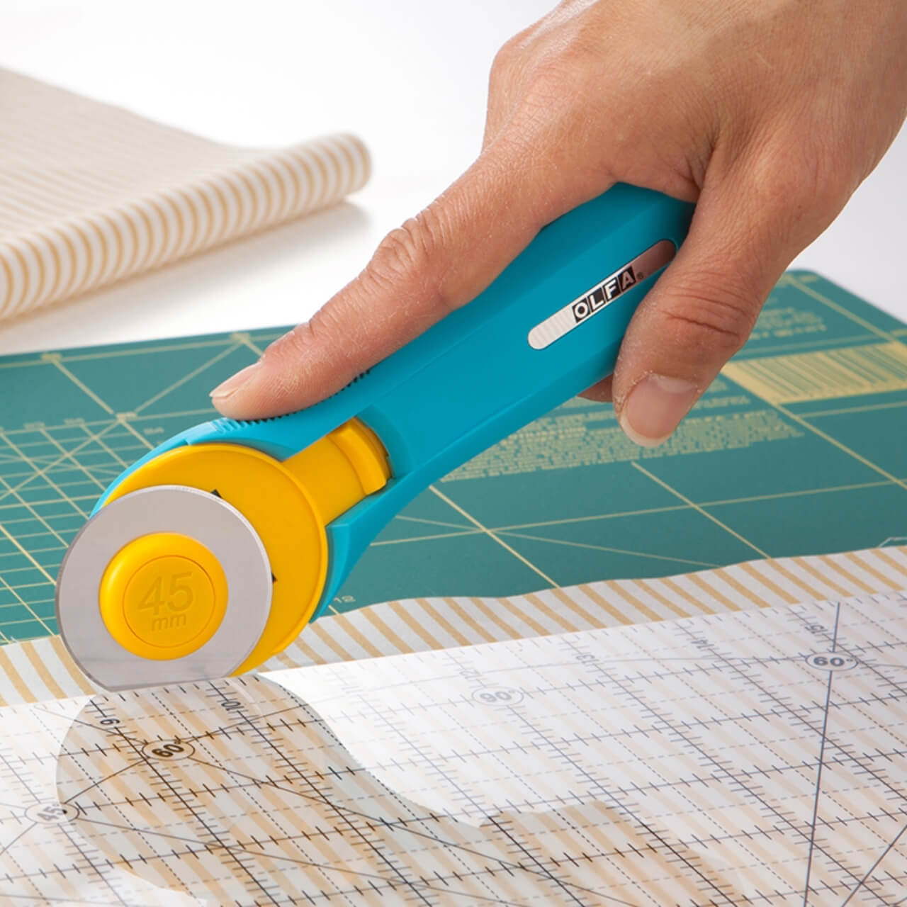 Close-up of a hand using the OLFA 45mm Quick-Change Rotary Cutter to cut through pale fabric, showing the cutter's blade and ergonomic aqua handle.