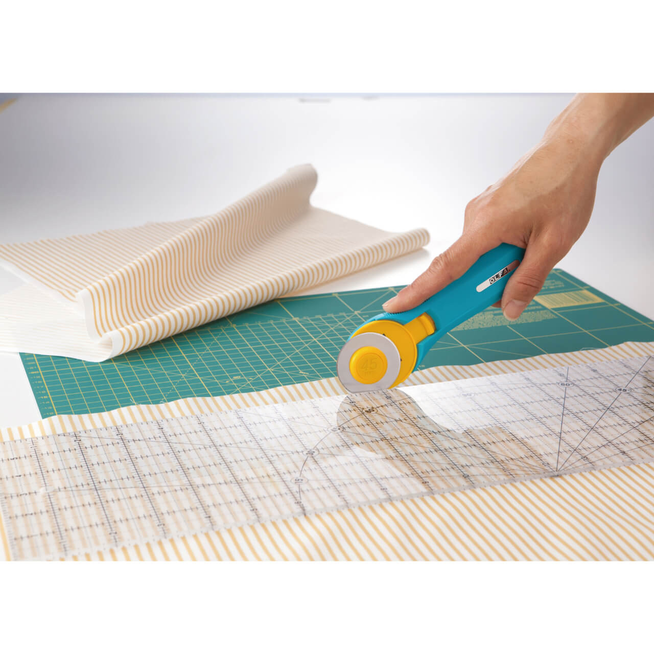 Hand using an OLFA 45mm rotary cutter alongside the OLFA 6 x 12-inch ruler to precisely cut striped fabric on a self-healing cutting mat