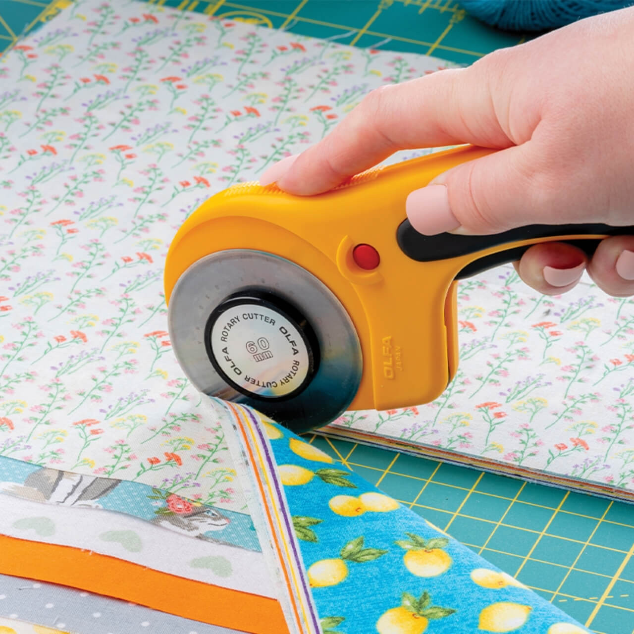 Close-up of a hand using the OLFA Deluxe Ergonomic mm Rotary Cutter to cut through multiple layers of fabric, showing the cutter's blade and ergonomic yellow handle.