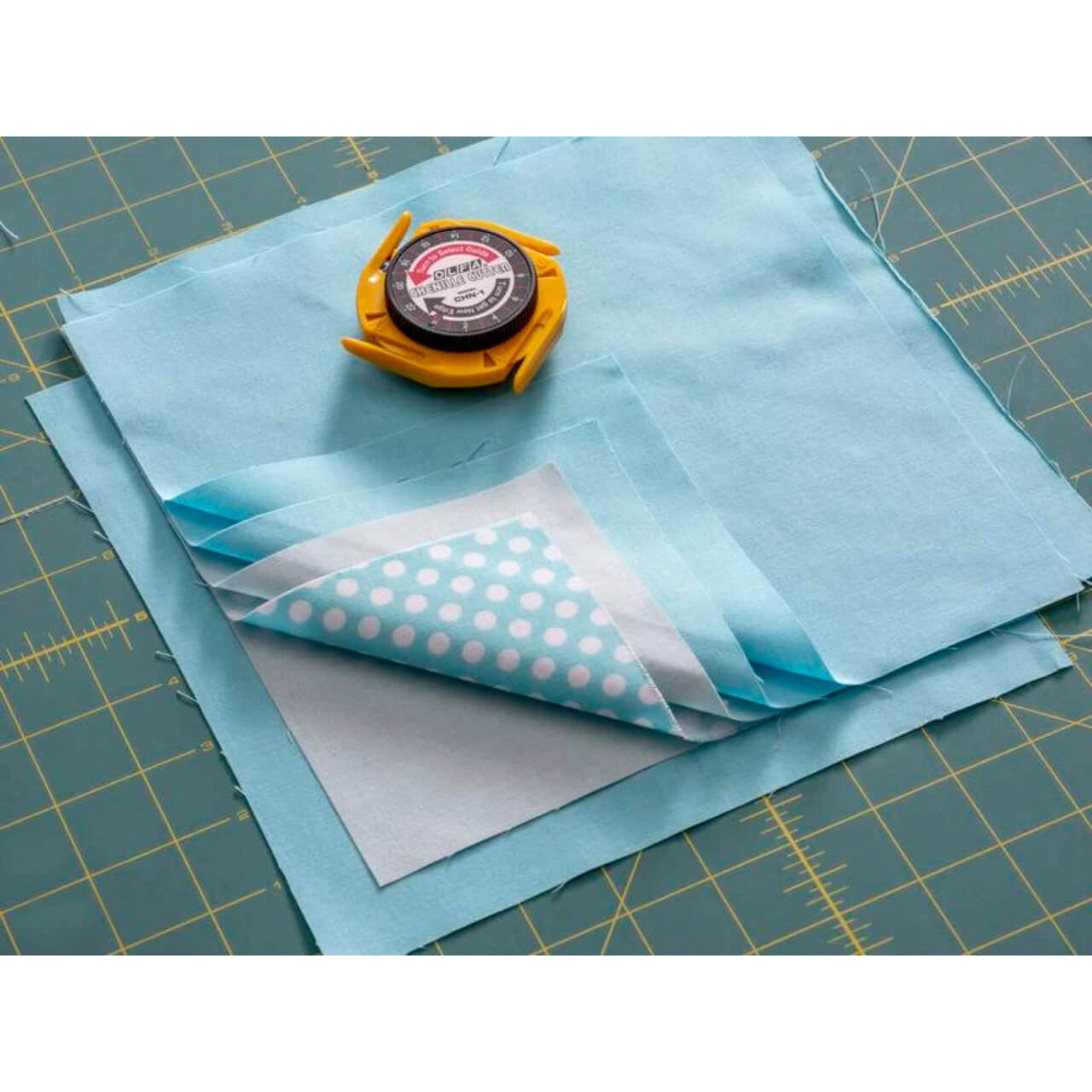 OLFA CHN-1 Chenille Cutter placed on a layered fabric of blue hues, ready for textile crafting on a cutting mat with grid lines.