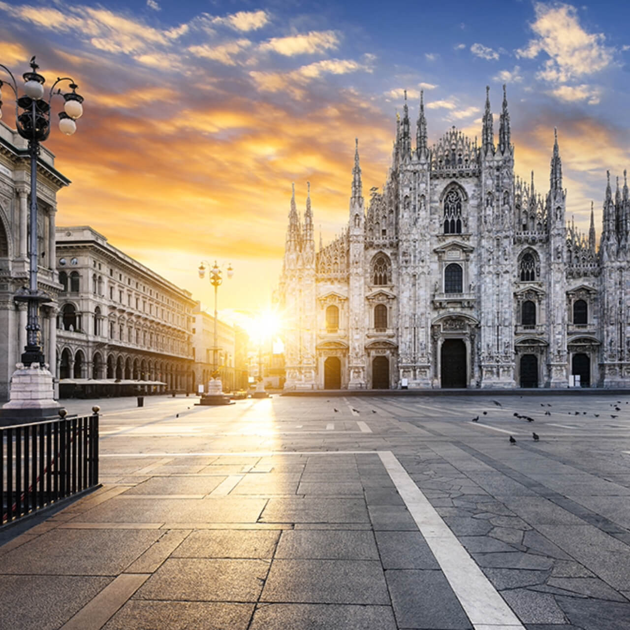 Sunrise over the Piazza del Duomo in Milan, with the sun casting a warm glow on the Gothic facade of the Duomo di Milano and the surrounding buildings, the square quiet and empty.