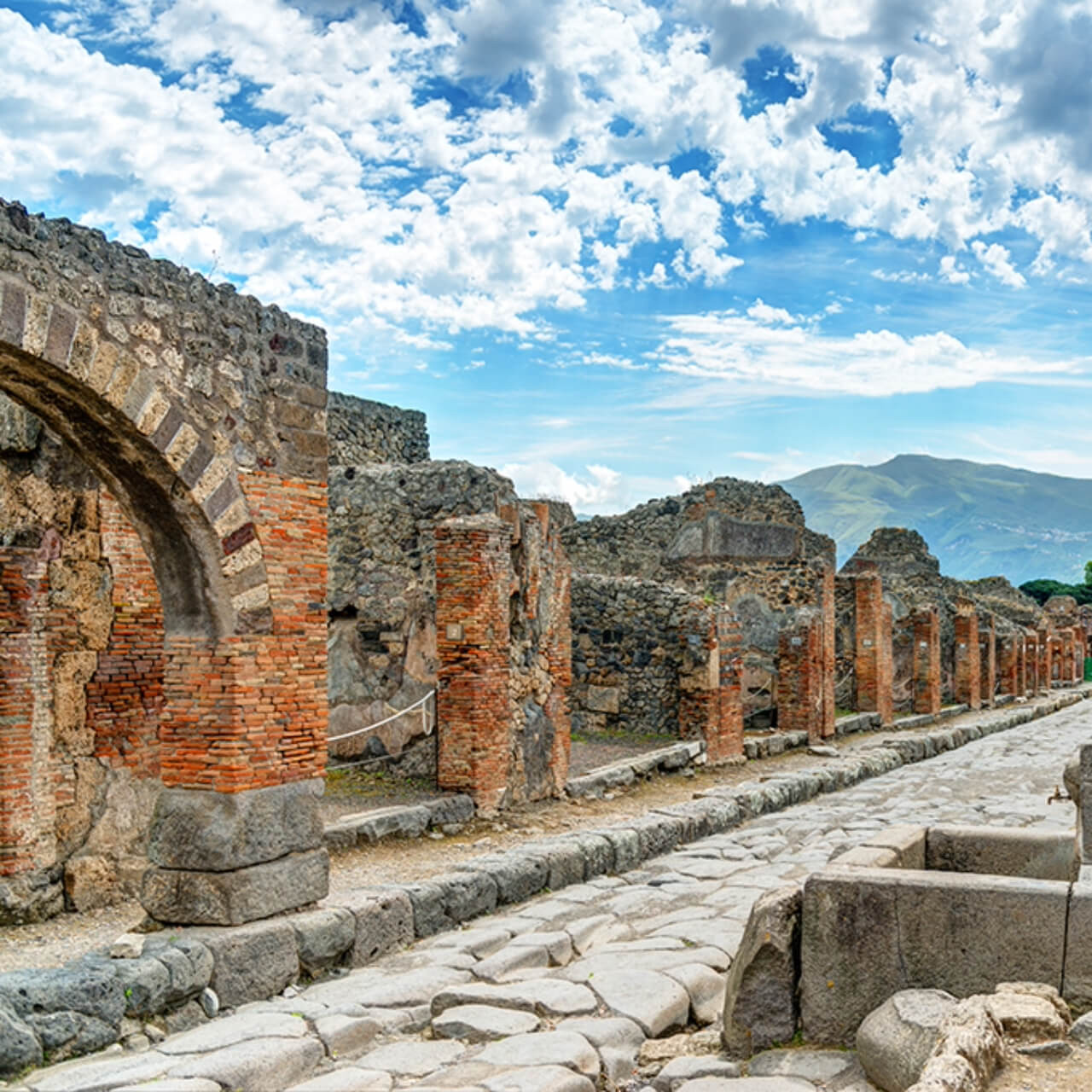 Ancient Roman ruins of Pompeii with cobblestone streets and remnants of red brick walls, with the silhouette of Mount Vesuvius in the background under a blue sky with scattered clouds.