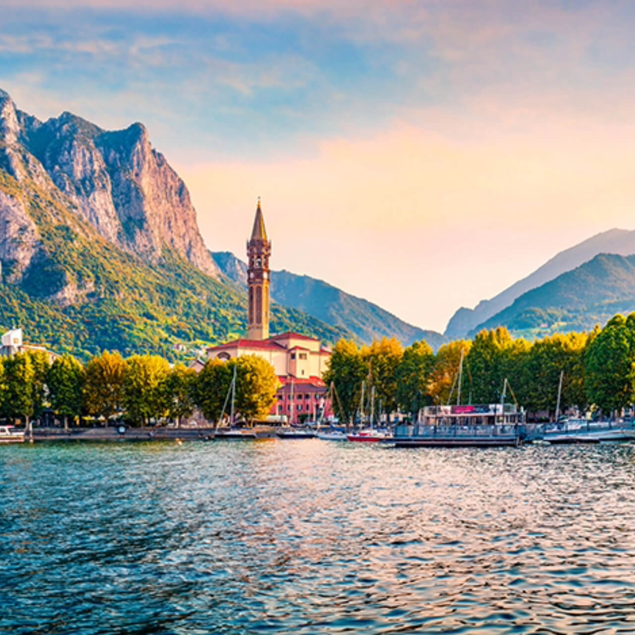 Picturesque view of Lake Como with a church spire rising against the backdrop of lush mountains and a serene lakefront lined with boats and trees under a soft dusk sky.