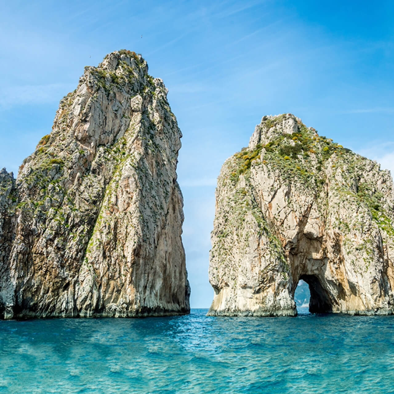 Majestic limestone cliffs of Capri with a natural arch over the azure waters of the Mediterranean Sea, under a bright blue sky.