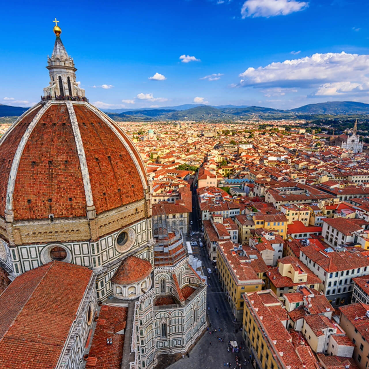 Aerial view of Florence with the iconic Duomo dominating the foreground, its red-tiled dome and intricate facade standing out against the cityscape and distant hills under a blue sky.
