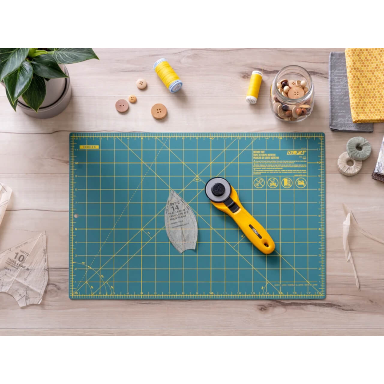 The image depicts a crafting scene on a wooden surface, featuring an OLFA green double-sided rotary mat with yellow grid lines and measurements. Centred on the mat is an OLFA rotary cutter with a vibrant yellow and black handle. The composition is enhanced by a patterned fabric piece, sewing threads in yellow and white, wooden buttons, and quilt patches, all neatly arranged around the mat, suggesting a peaceful quilting or crafting session.