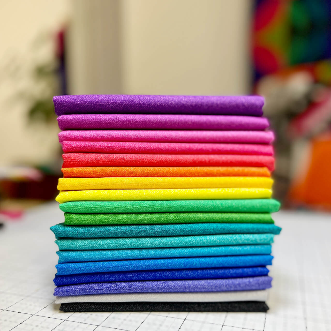 Stack of Phosphor Electric fat quarters in vibrant gradient colours on a sewing grid table.