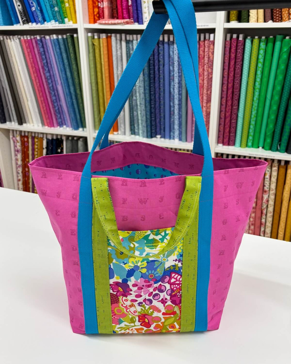 Bond Street Tote sewn in bright Alison Glass fabrics—pink body, blue long straps, green short handles, and a colourful feature pocket—shown in the quilt shop.