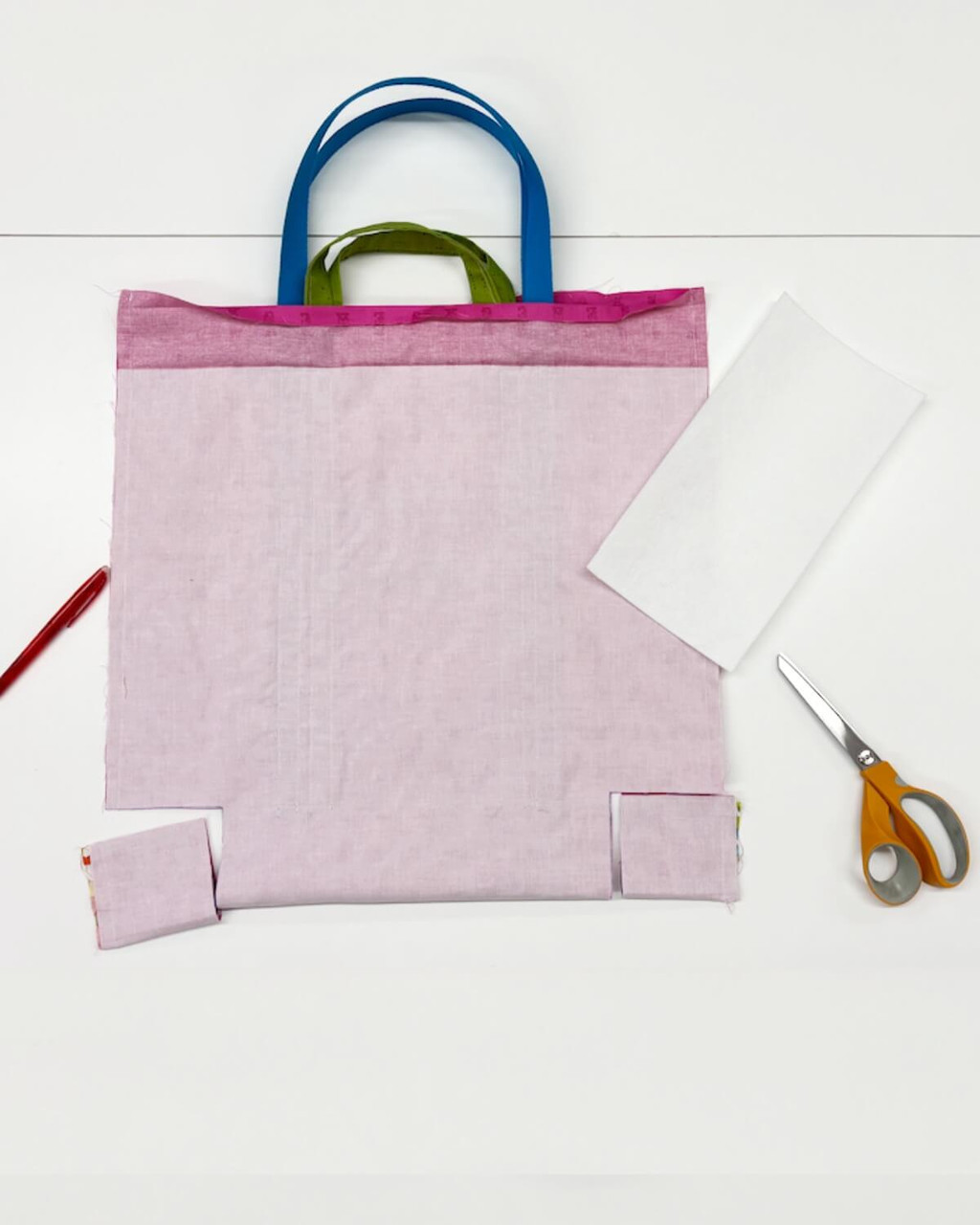 Wrong side of the tote body laid flat with short and long handles shown at the top. Marked top hem, square cut-outs at both lower corners for boxed corners, interfacing piece to the side, red fabric marker and scissors visible.