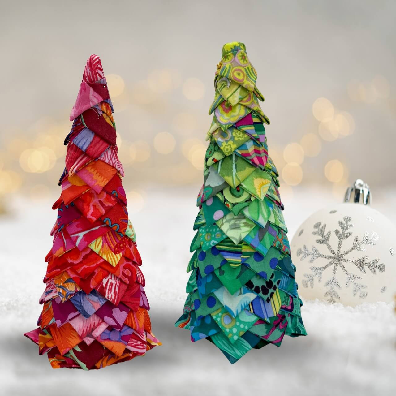 Two handmade no-sew Christmas trees crafted from Kaffe Fassett fabrics, one in red tones and one in green, displayed on a snowy background with festive baubles.