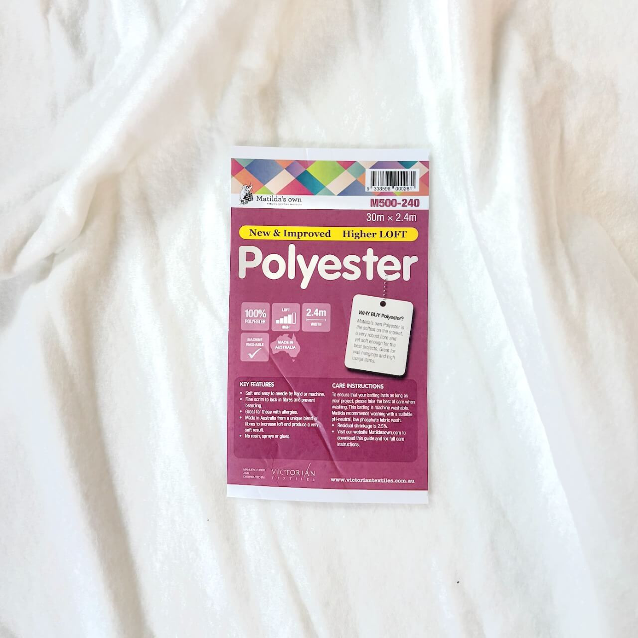 A close-up of Matilda's Own 100% Poly Quilt Wadding with its pink product label placed on top. The label highlights key features" It also mentions a quilting distance of "8-10 inches" and that the wadding is "Made in Australia." A section on the label explains the benefits of using polyester wadding, emphasising its softness and durability. Care instructions and key product details are listed at the bottom, along with the Victorian Textiles logo and website information. The wadding itself appears soft, white and a slightly textured surface. A close-up of Matilda's Own 100% Poly Quilt Wadding with its pink product label placed on top. The label highlights key features" It also mentions a quilting distance of "8-10 inches" and that the wadding is "Made in Australia." A section on the label explains the benefits of using polyester wadding, emphasising its softness and durability. Care instructions and key product details are listed at the bottom, along with the Victorian Textiles logo and website information. The wadding itself appears soft, white and a slightly textured surface.