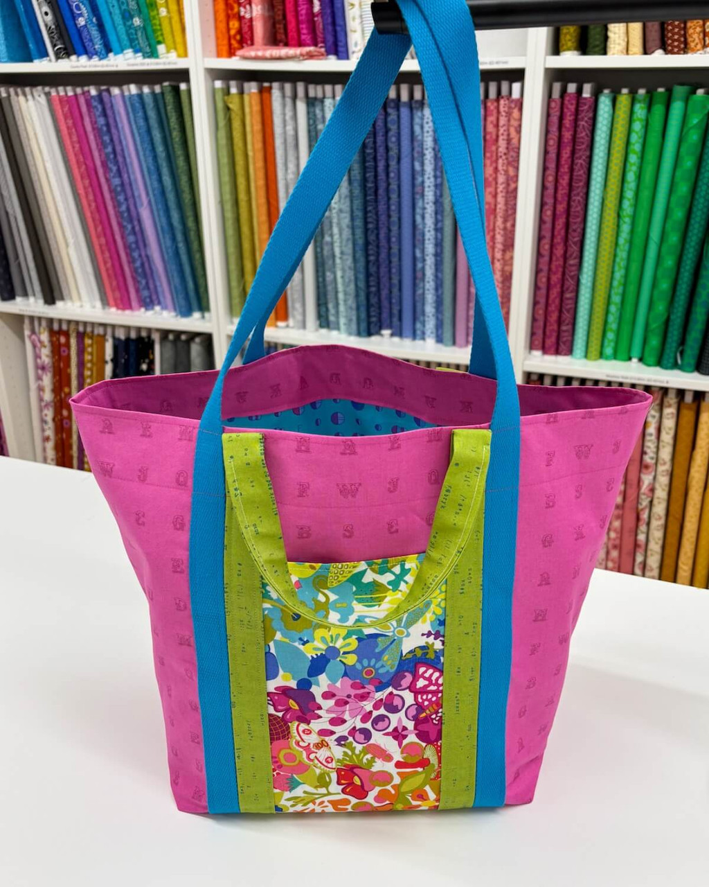 Bond Street Tote sewn in bright Alison Glass fabrics—pink body, blue long straps, green short handles, and a colourful feature pocket—shown in the quilt shop.
