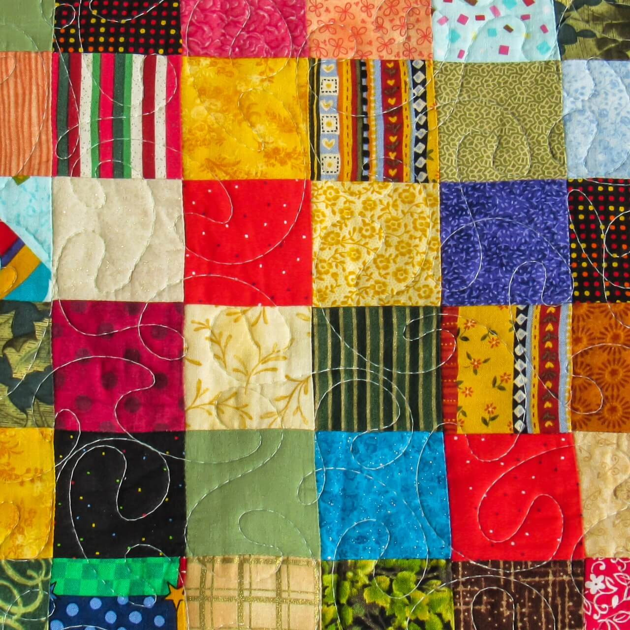 Colourful patchwork quilt made from small fabric squares, quilted with flowing free motion quilting lines stitched across the surface.