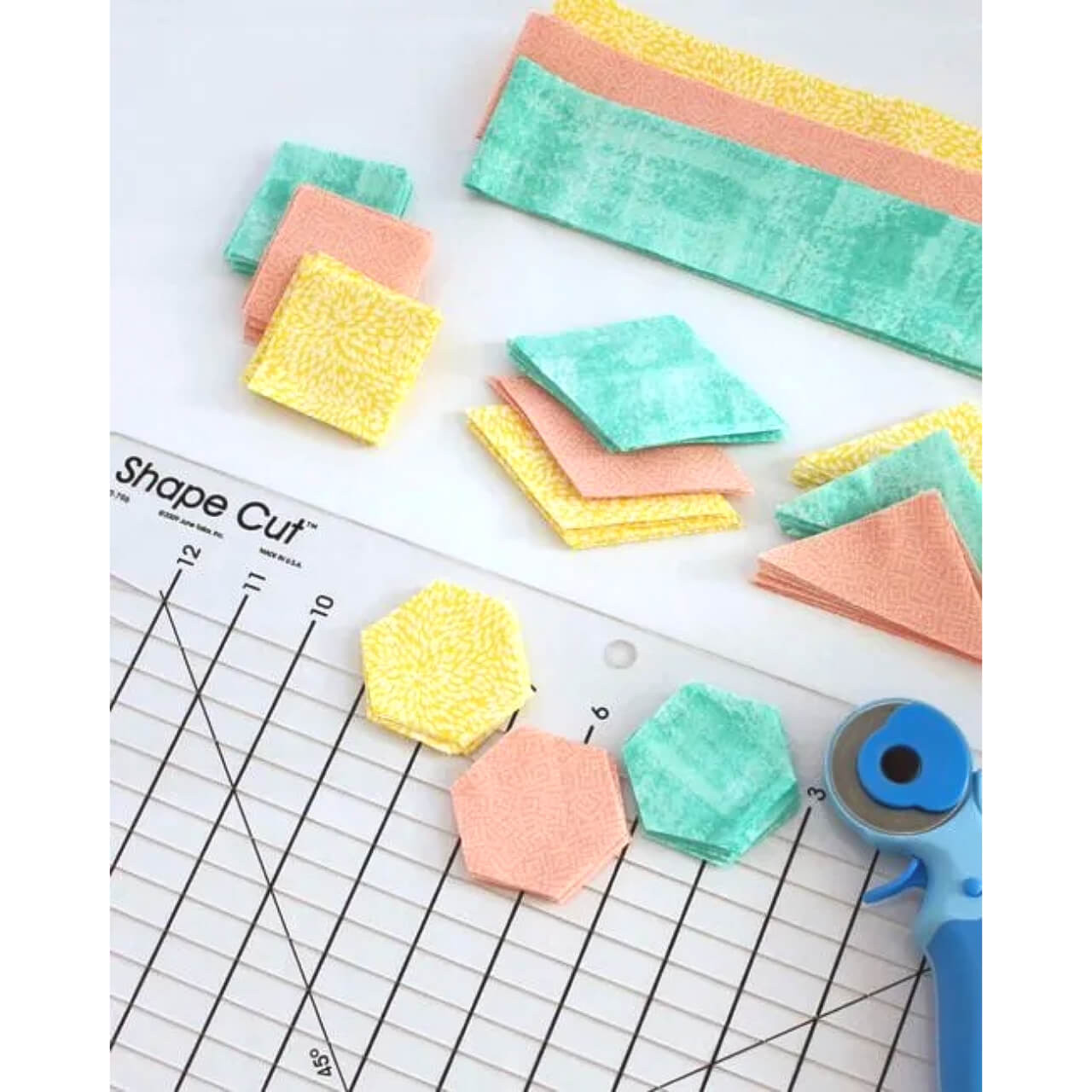 A selection of neatly cut fabric squares and hexagons in yellow, aqua, and peach colors arranged next to the June Tailor Shape Cut Ruler, with a rotary cutter, showcasing the ruler’s ability to cut multiple shapes for patchwork projects.