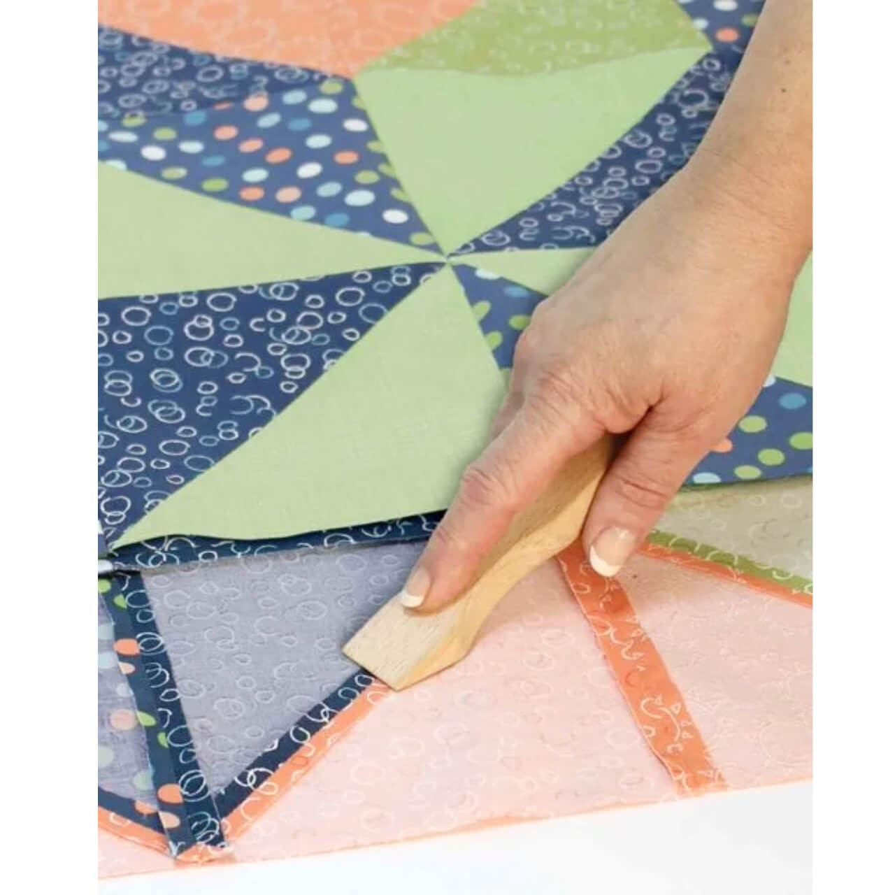 A hand using the June Tailor Magic Seam Wand to press and flatten the seams of a colorful patchwork quilt, demonstrating the tool's effectiveness in creating crisp fabric seams without the use of an iron.