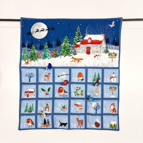 Quilted blue Advent calendar panel featuring a snowy cottage scene, woodland animals, Santa’s sleigh in the night sky, and 24 numbered pockets for Christmas surprises.