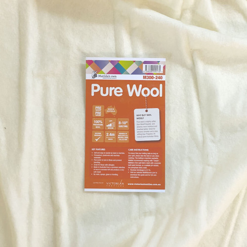 A close-up of Matilda's Own 100% Wool Quilt Wadding with its orange product label placed on top. The label highlights key features, including "100% Preshrunk Wool," "Machine Washable," and "Prewashed & Preshrunk." It also mentions a quilting distance of "8-10 inches" and that the wadding is "Made in Australia." A section on the label explains the benefits of using pure wool wadding, emphasising its softness, moisture-wicking properties, and breathability. Care instructions and key product details are listed at the bottom, along with the Victorian Textiles logo and website information. The wadding itself appears soft, with a natural off-white hue and a slightly textured surface.