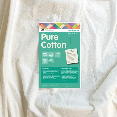 A close-up of Matilda's Own extra wide 100% Cotton Quilt Wadding with its green product label placed on top. The label highlights key features, including "100% Cotton," "Machine Washable," and "2.4m Width." It also states that the wadding is "Made in Australia" and provides a low loft for a traditional quilting finish. A section on the label explains the benefits of pure cotton wadding, mentioning its breathability, softness, and suitability for heirloom quilting. Care instructions and key product details are listed at the bottom, along with the Victorian Textiles logo and website information. The wadding itself appears soft, off-white, and textured.