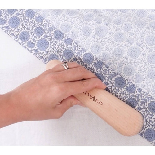 The Milward Tailor's Clapper in use, pressed onto a folded seam on blue floral quilting fabric, with the Milward branding visible on the wood.