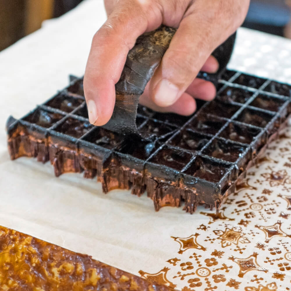 A person's hand pressing a traditional batik wax stamp onto fabric, creating intricate patterns.