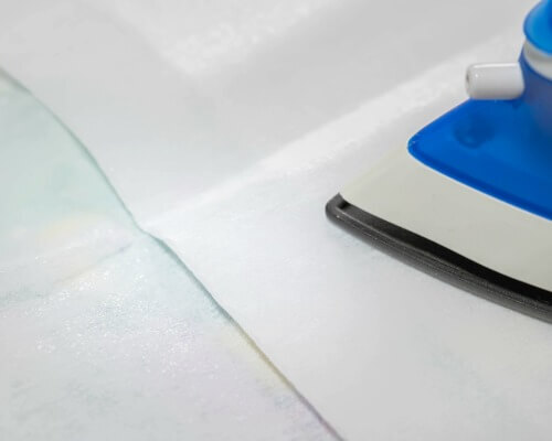 Dry iron pressing a paper-backed fusible web sheet onto fabric to create a permanent bond.