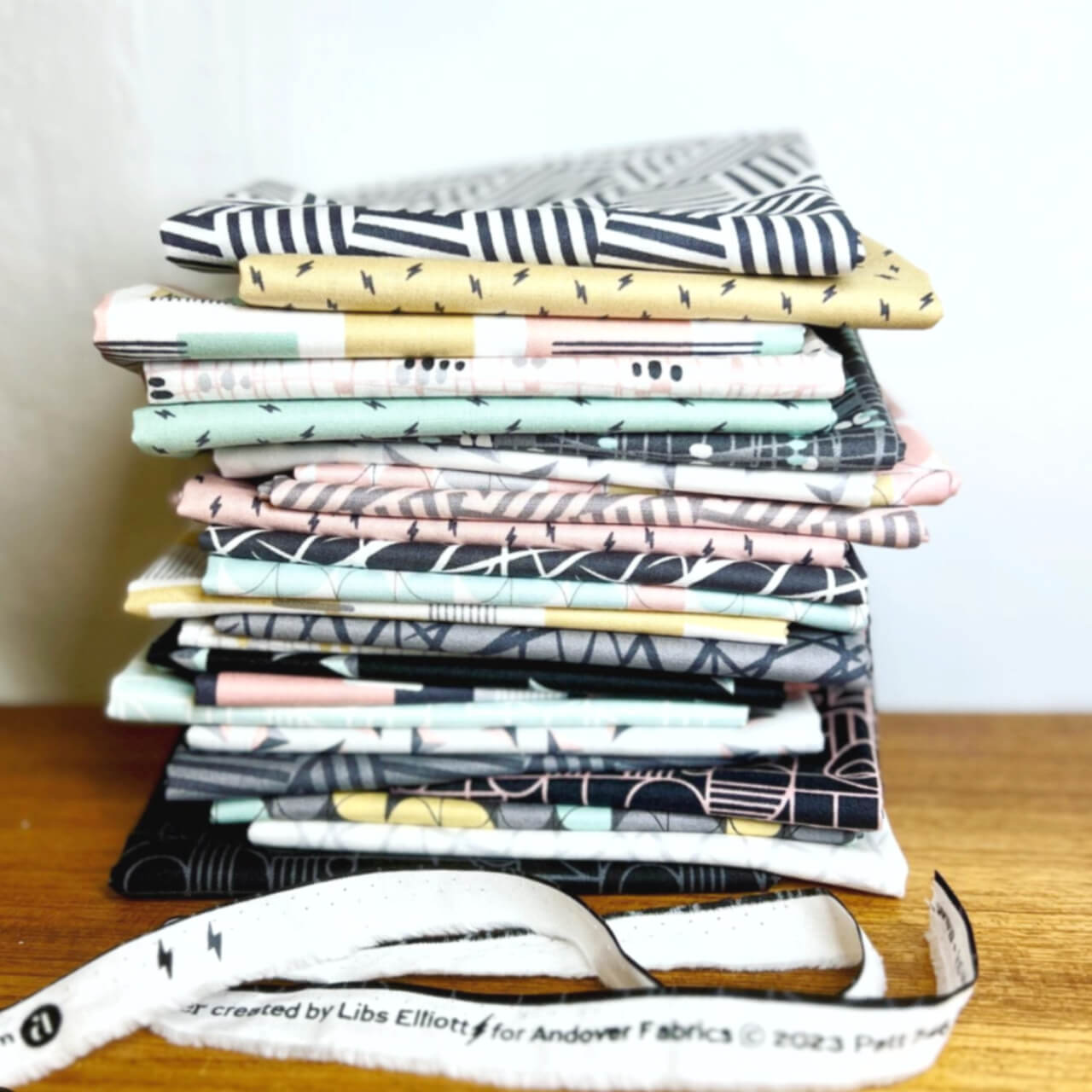 Stack of Rancho Relaxo fat quarter fabrics by Libs Elliott for Andover Fabrics, showcasing varied mid-century inspired geometric patterns in a palette of black, white, pink, yellow, and aqua. The pile of fabric is loosely stacked on a wooden table.