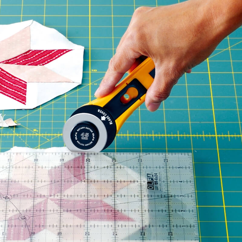 Hand using an OLFA rotary cutter on a quilt patchwork block with a cutting mat and ruler.