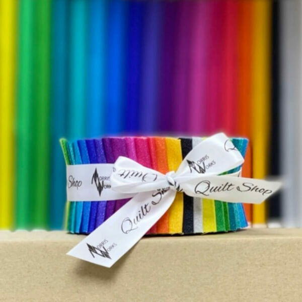 Vibrantly coloured 2.5 inch Jelly Roll fabric strips tied with a ribbon, displayed against a colourful fabric shelf background.