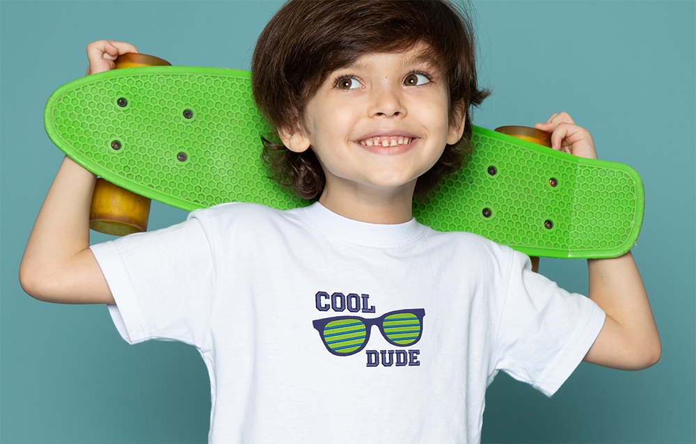 Boy holding a skateboard, wearing a white t-shirt with 'Cool Dude' and sunglasses embroidered on the front.