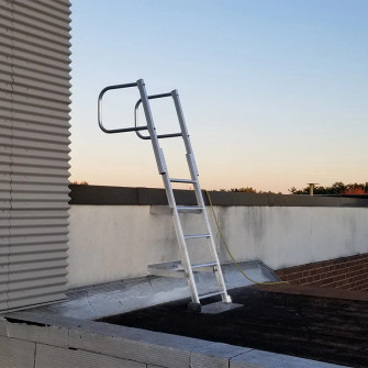Aluminum fixed access ladder with a safety hoop mounted on a rooftop wall at dusk