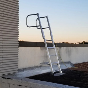 Aluminum fixed access ladder with a safety hoop mounted on a rooftop wall at dusk