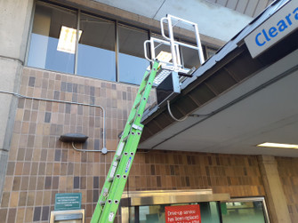 Green extension ladder resting against a white KD001 ladder dock landing platform mounted at a commercial roof edge