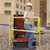 Woman in a hard hat climbing through an open roof hatch with yellow safety railing, city skyline behind