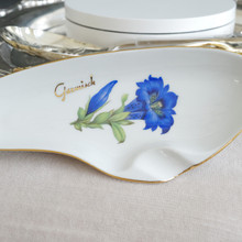 Close up view of a vintage white porcelain ashtray with blue flower, gold rim, and gold Garmisch in interesting organic oval shape that nods to a MCM mid-century modern style.