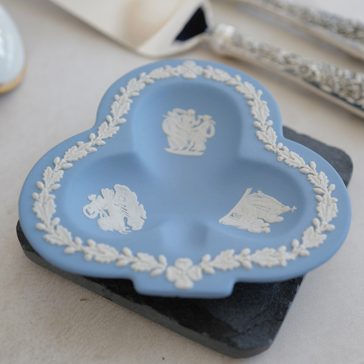 Vintage Wedgwood Jasperware ring dish in a unique club shape, crafted in England. Featuring a pale blue (lavender) background with intricate white relief designs of classical figures, flowers, and leaves. Perfect as a jewelry or trinket dish, this 4.5" x 4.5" collector's piece is in excellent condition with no cracks or chips.