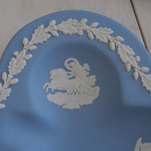 This is a close up of a vintage ring dish is in a club shape and features a classical design in white against the light blue background.  In the center of the each recess, there is a relief of figures.