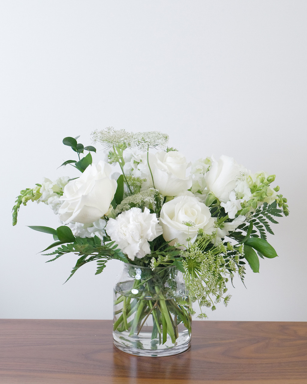 In Loving Memory sympathy bouquet with white roses, snapdragons or stock, dreamy white blooms and greenery, thoughtfully arranged in a clear glass vase.