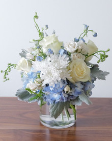 Lake Effect arrangement of icy blue hydrangea, white roses, and cream spray roses in a clear glass vase, light blue delphinium drifting out the top