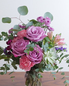 The Keeper arrangement on a table in natural light, pink and lavender blooms with silver dollar eucalyptus and sword fern in a glass vase.