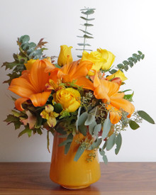A vibrant autumnal arrangement features golden yellows, oranges, and fall leaves.  Designed in a orange glass vase.