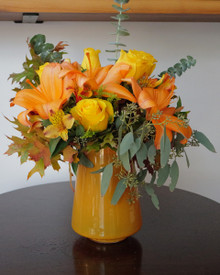 Fall flower arrangement of orange lilies, yellow roses, eucalyptus, and fall leaves in an orange glass vase.
