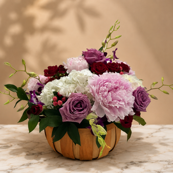 Elegant floral centerpiece featuring blush pink peonies, lavender roses, red roses, white hydrangea, and orchids arranged in a natural wooden bowl on a marble surface with a warm neutral background.