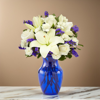 Standard Elegant white lilies, roses, and carnations accented with deep purple statice, beautifully arranged in a vibrant cobalt blue glass vase. This classic sympathy and remembrance bouquet conveys peace, serenity, and heartfelt condolences, perfect for funeral services or thoughtful home displays.