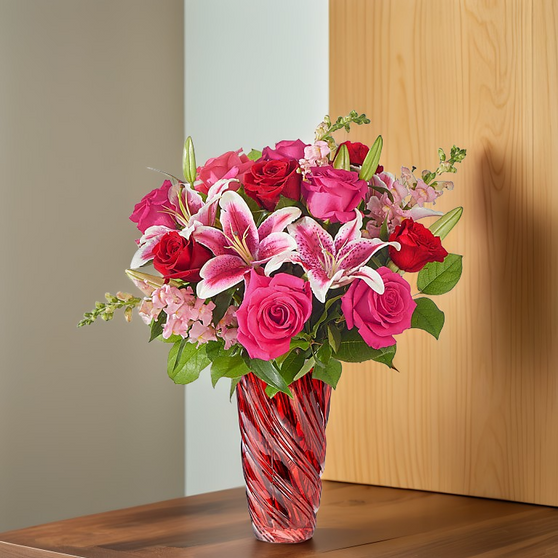 Compact bouquet of pink roses, red roses, and pink stargazer lilies arranged in a red swirl glass vase, set on a table with a light wood background.